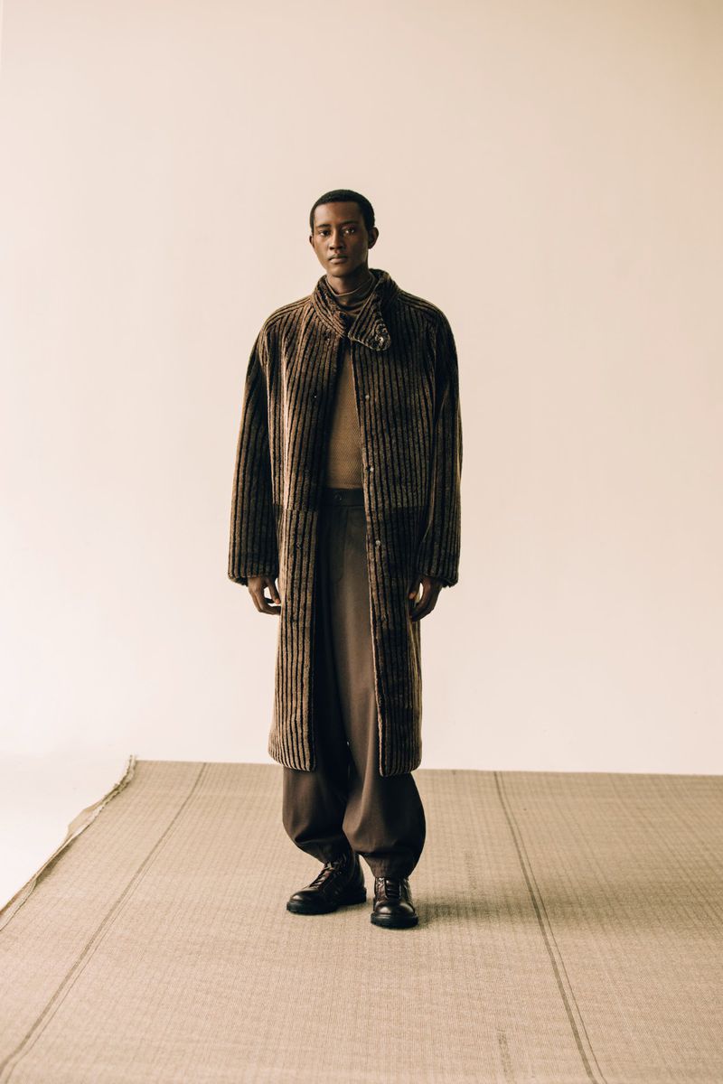 Full-length fashion portrait of an adult Black male model wearing a brown striped coat, brown pants, and black leather shoes in a studio.