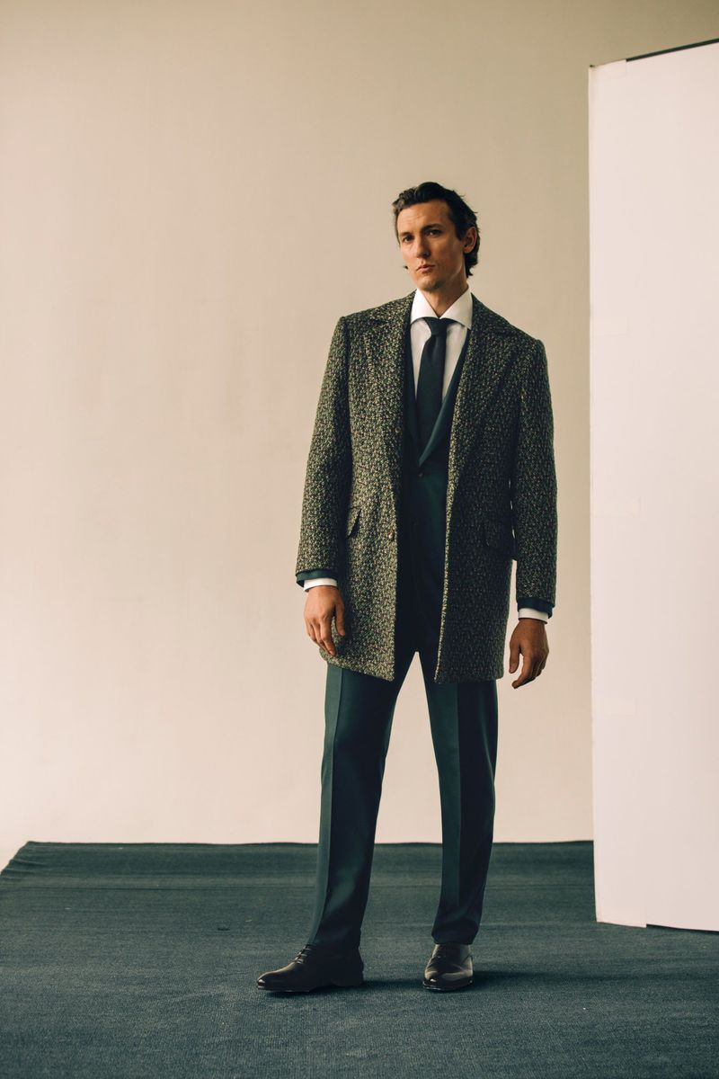 A man in a dark green suit, tie, and brown patterned overcoat strikes a confident pose against a minimalist backdrop in a fashion editorial.