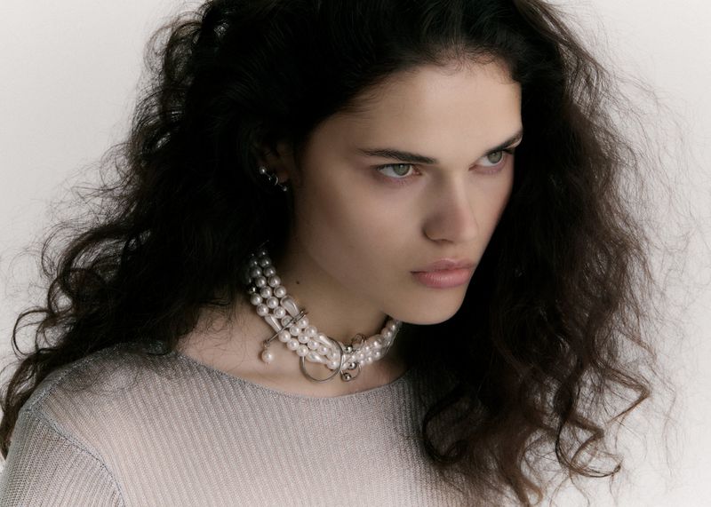 Close-up studio shot of an adult woman with long, dark wavy hair, wearing a layered pearl necklace and a light-colored top.