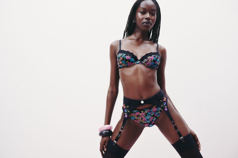 A Black woman poses in floral lingerie and thigh-high stockings against a light backdrop in a studio shot, showcasing a confident and elegant look.
