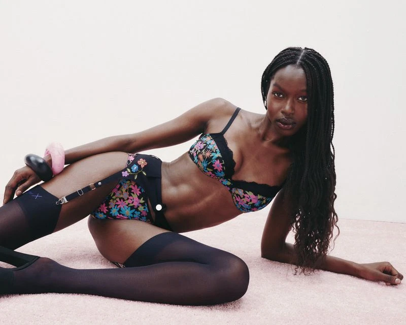 A black model poses in floral lingerie set with black stockings and high heels on a pink carpet against a white wall in a studio.