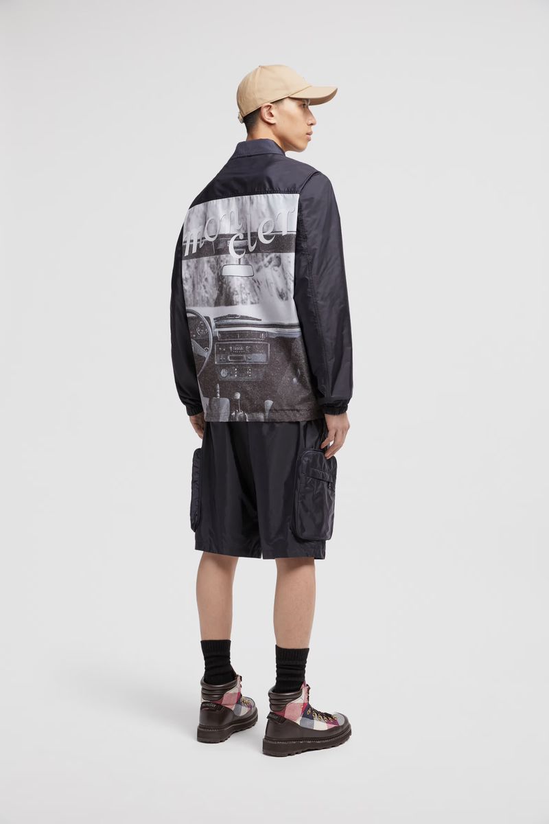 Back view of a slender adult male wearing a dark jacket with an image on the back, paired with cargo shorts and boots.