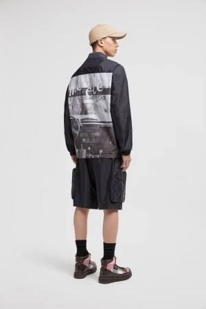 Back view of a slender adult male wearing a dark jacket with an image on the back, paired with cargo shorts and boots.