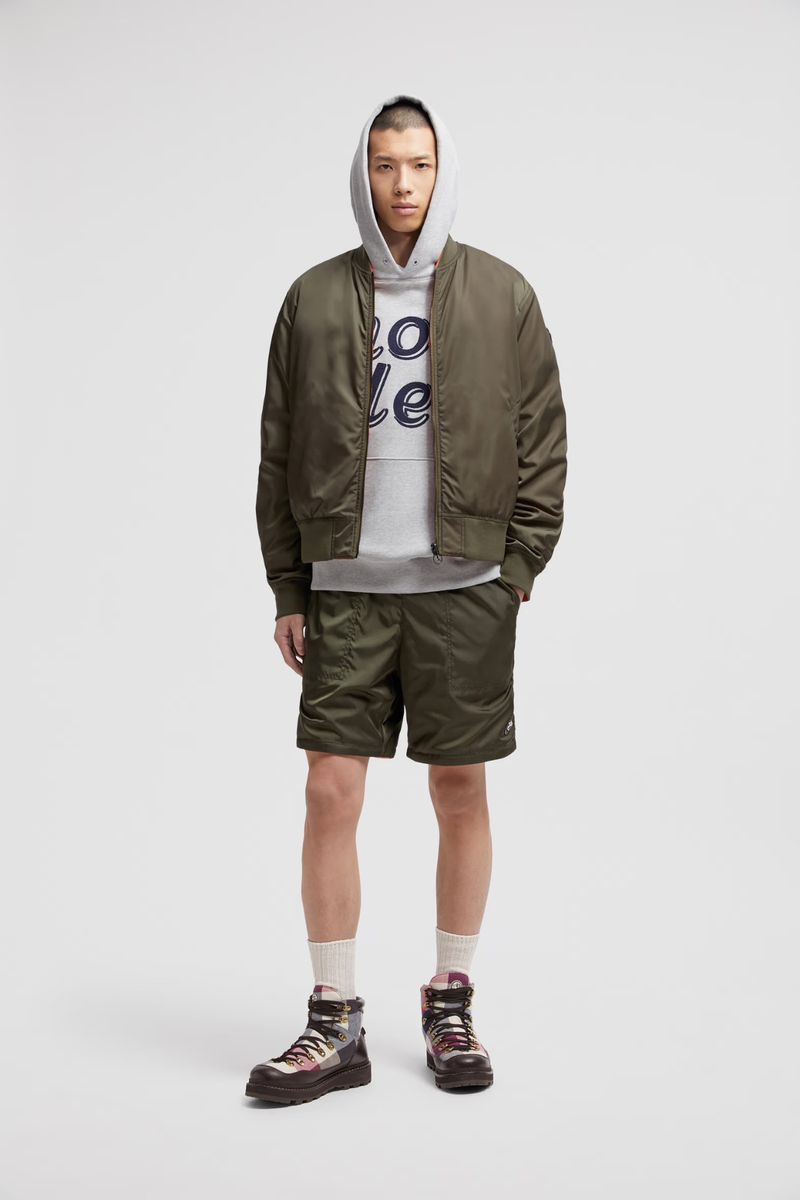 Full-length shot of a male model in an olive green bomber jacket and matching shorts, posing against a neutral backdrop in a studio.