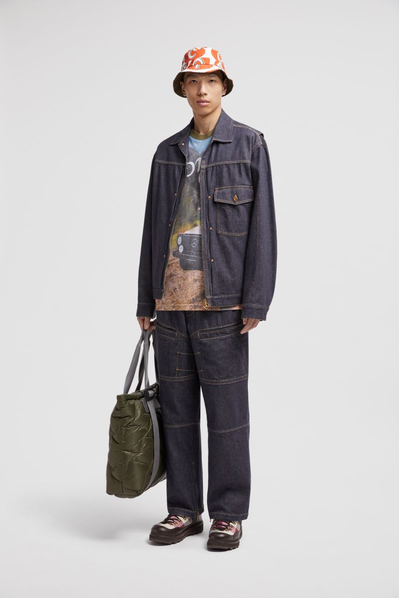 Asian man poses in dark denim jacket, pants, floral bucket hat, holding green duffel bag on neutral background in a studio shot.