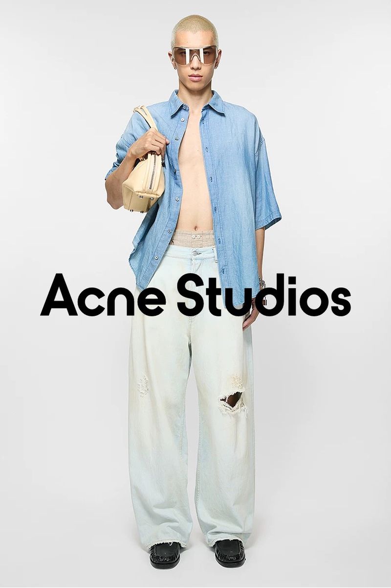 Male fashion model poses in unbuttoned light blue shirt, baggy pants, and sunglasses, holding purse, in a white photo studio setting.