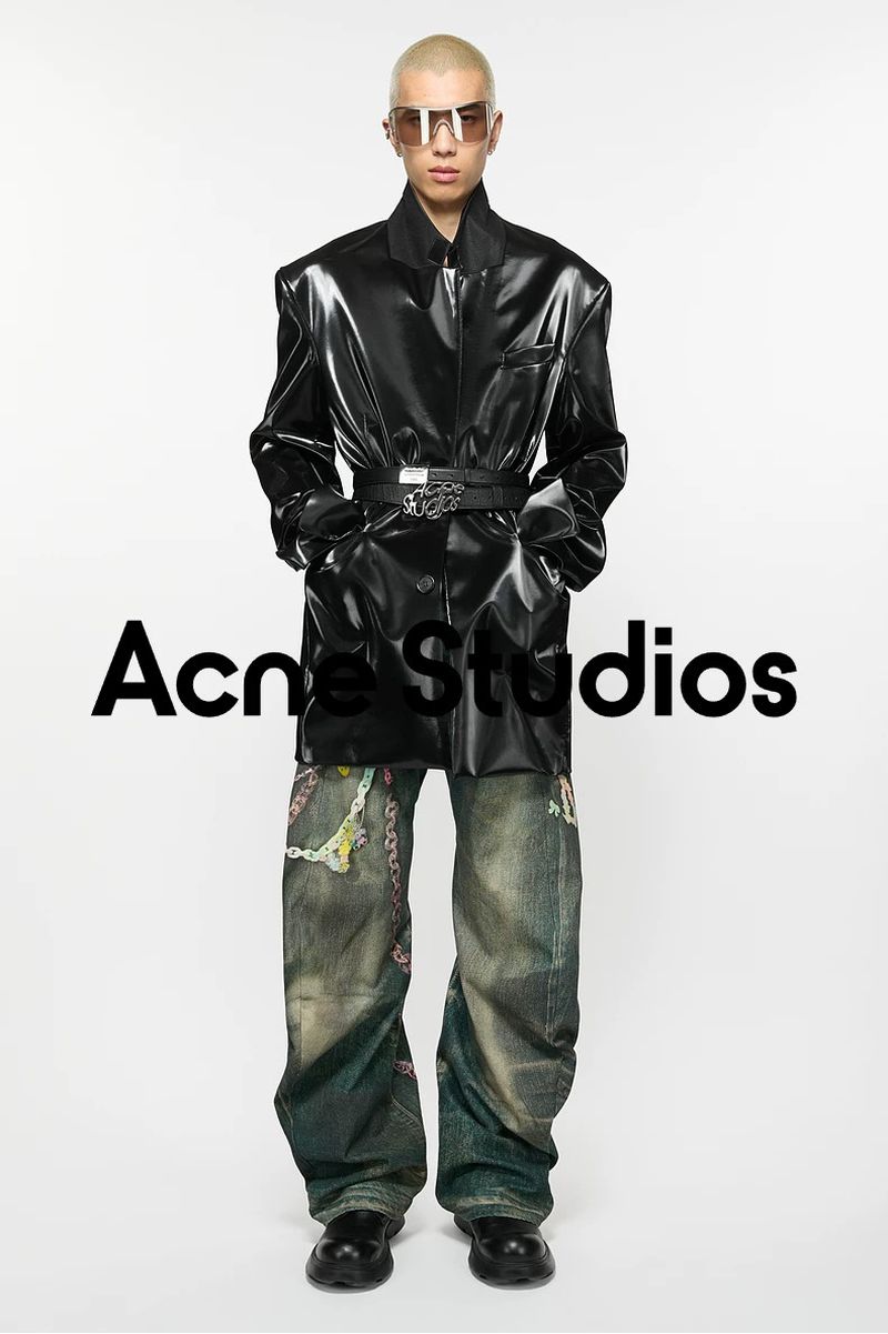 Bald male model posing in a black shiny trench coat, belt, patterned jeans, and black shoes in a studio, showcasing a fashion brand.