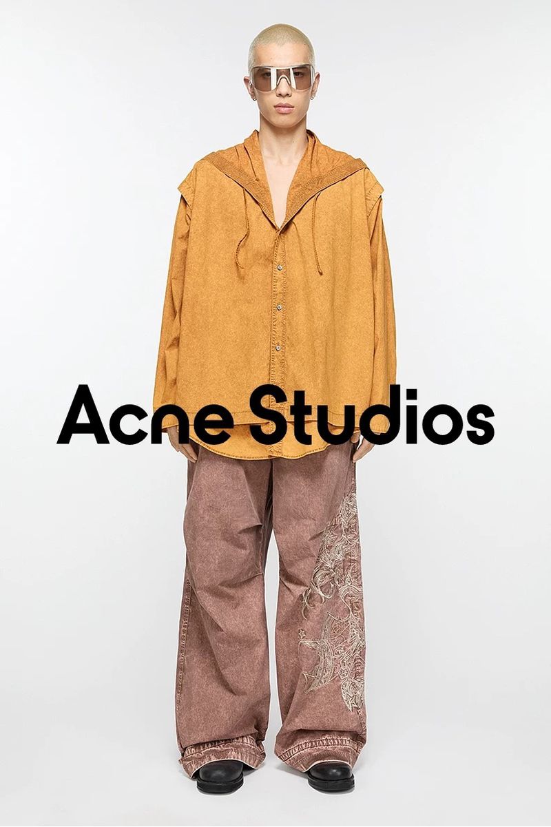 An adult fashion model poses in a mustard hooded shirt and rose-colored pants with embroidery against a plain white backdrop.