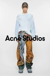 Model wearing a textured light blue sweater, patterned trousers, and a cap against a white background.