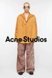 Model wearing tinted sunglasses, an orange hooded shirt, and wide printed trousers against a plain white background.