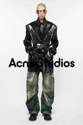 Model with shaved blonde hair wearing shiny black jacket, tinted sunglasses, and distressed printed jeans on white background.