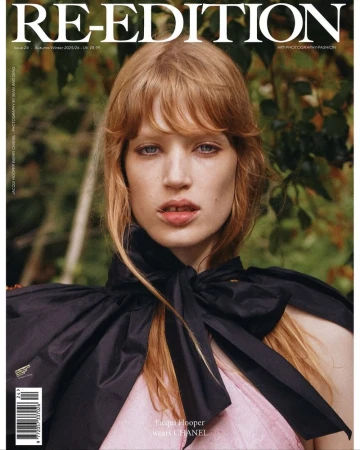 Portrait of a model with reddish blonde hair wearing a black bow blouse outdoors against foliage.