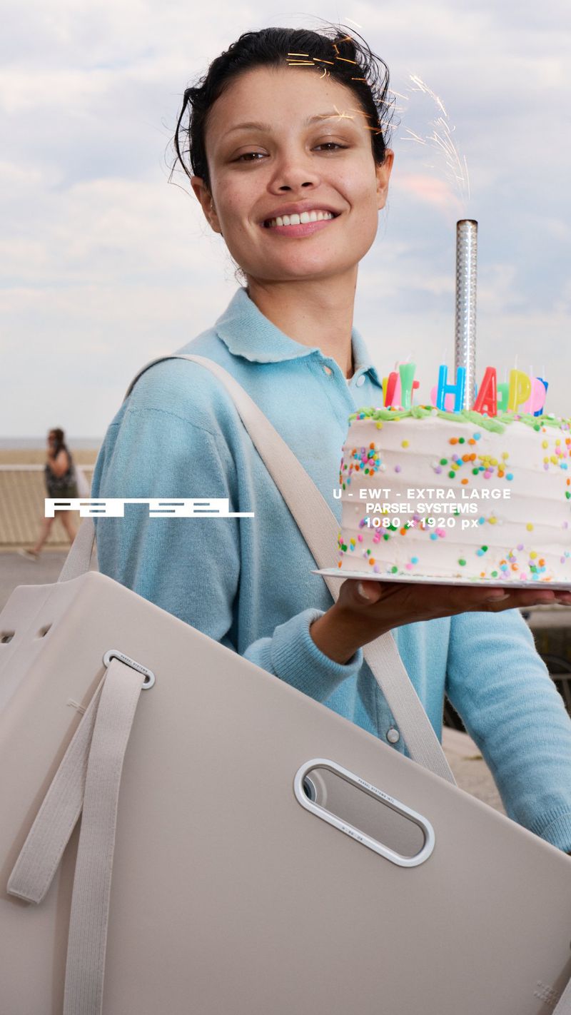 Woman holding a birthday cake on the beach with sparklers, wearing a light blue shirt and carrying a large tote bag over her shoulder.