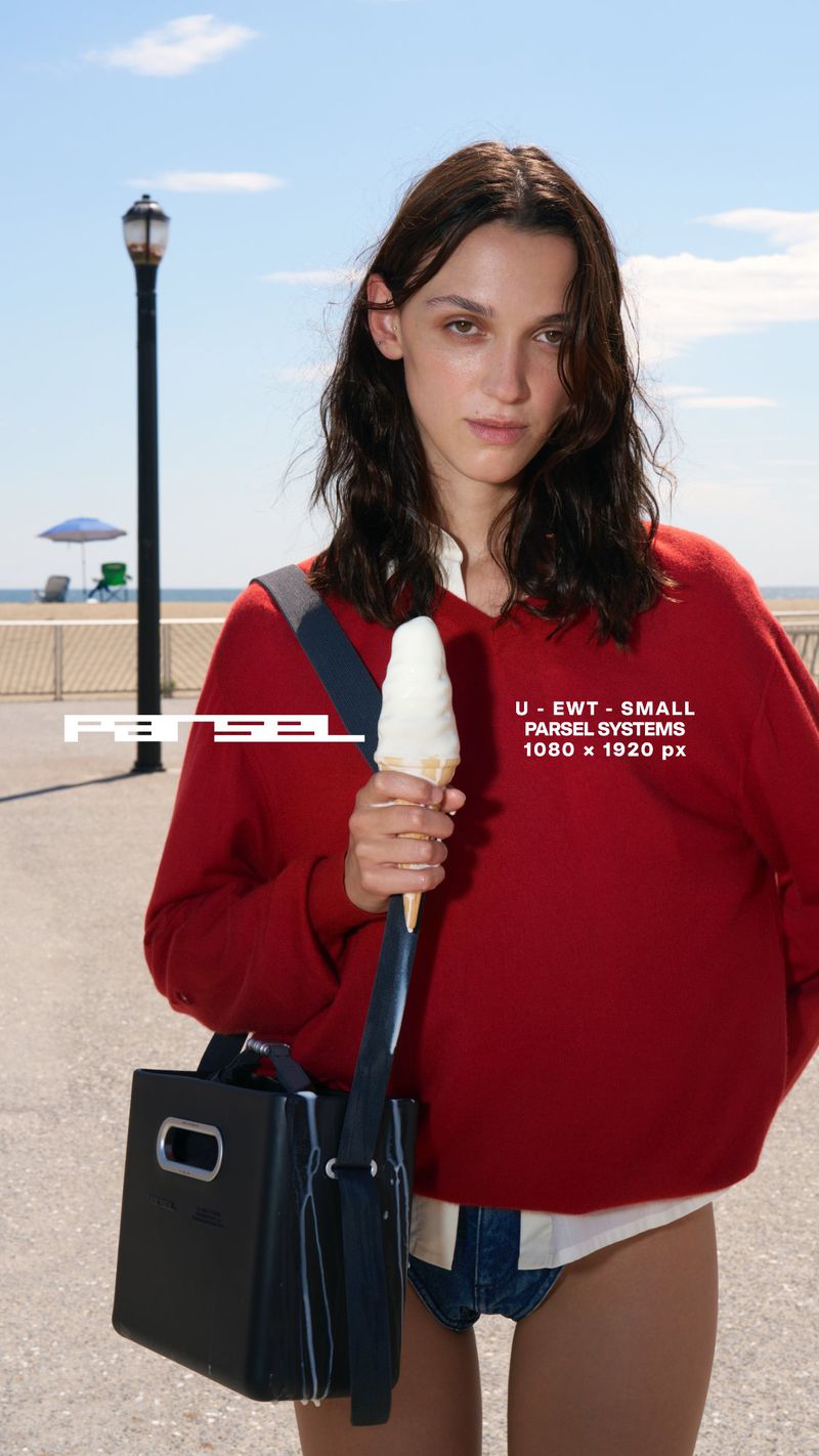 Woman in red sweater holds ice cream and black satchel on boardwalk near beach under blue sky on sunny day. She gazes at the viewer.