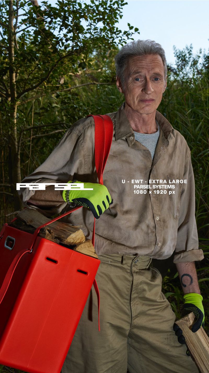Mature man carries a red container filled with firewood, wearing work gloves and a tan-colored shirt, in an outdoor wooded environment.