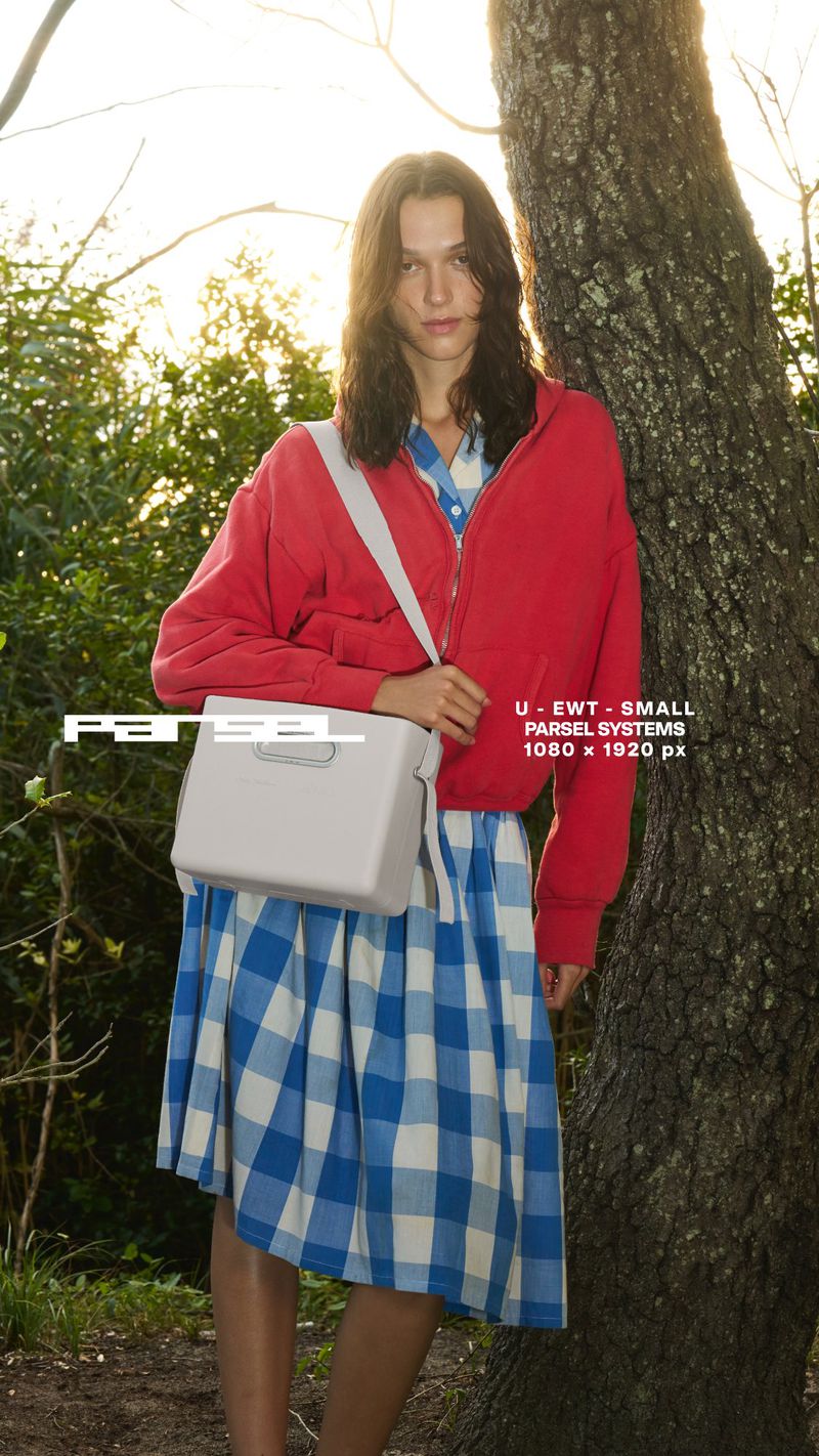 An adult female model poses outdoors, wearing a red jacket, blue and white gingham dress, and a white purse in natural lighting.