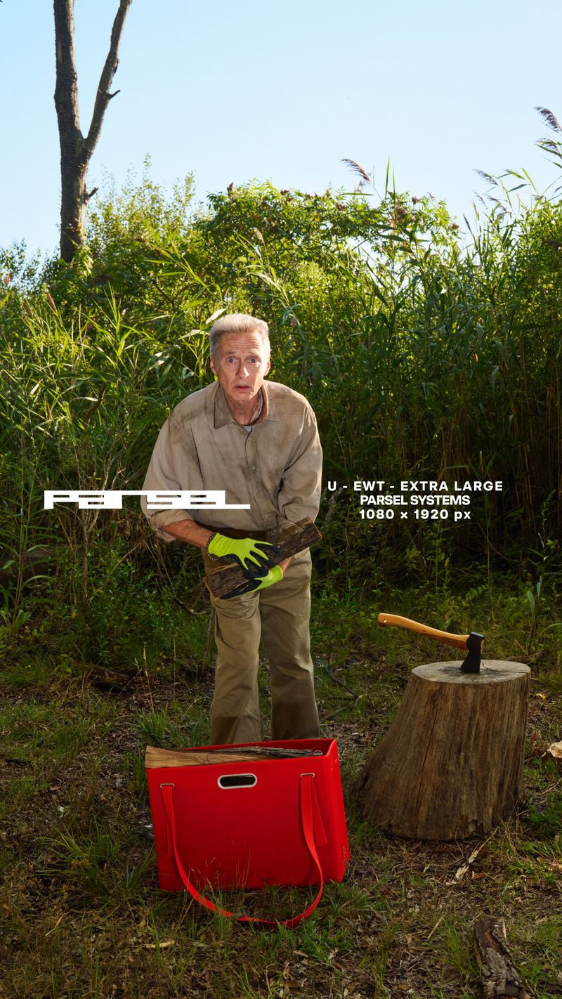 Older man chopping wood outdoors with an axe, wearing work gloves. A red bag with firewood sits near the stump.