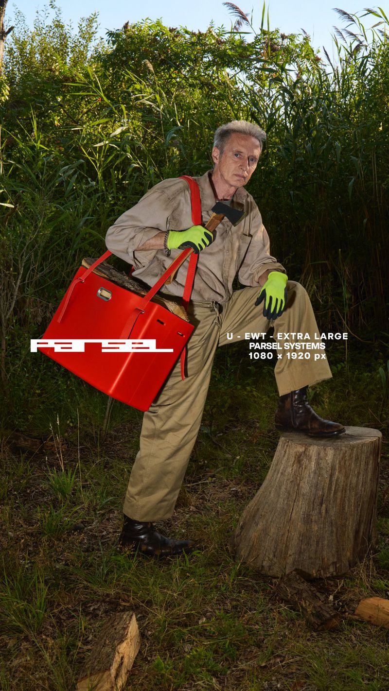 Adult male outdoors wearing coveralls, boots, and gloves, carrying a red tote bag and holding an axe, posing on a tree stump.