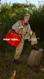 An older man wearing workwear holding a bright red bag while posing outdoors next to a tree stump.