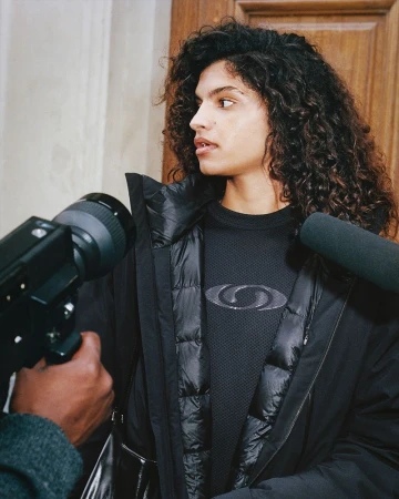 Woman with curly hair poses in black jacket as someone holds a camera up to her face indoors with neutral wall and wood door.