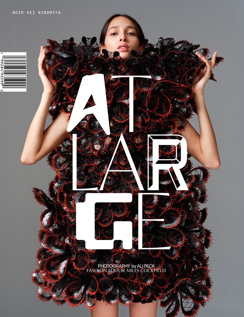 Fashion editorial shot of an adult female model wearing a black sculptural dress standing in front of a grey studio background with text overlay.