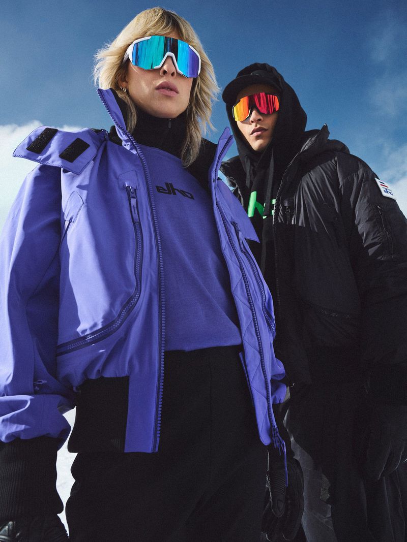 Two people wearing ski goggles and winter jackets pose against a cloudy sky. One person wears a purple jacket, the other wears a black one.