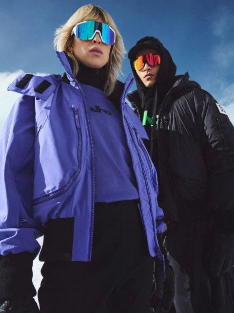 Two people wearing ski goggles and winter jackets pose against a cloudy sky. One person wears a purple jacket, the other wears a black one.