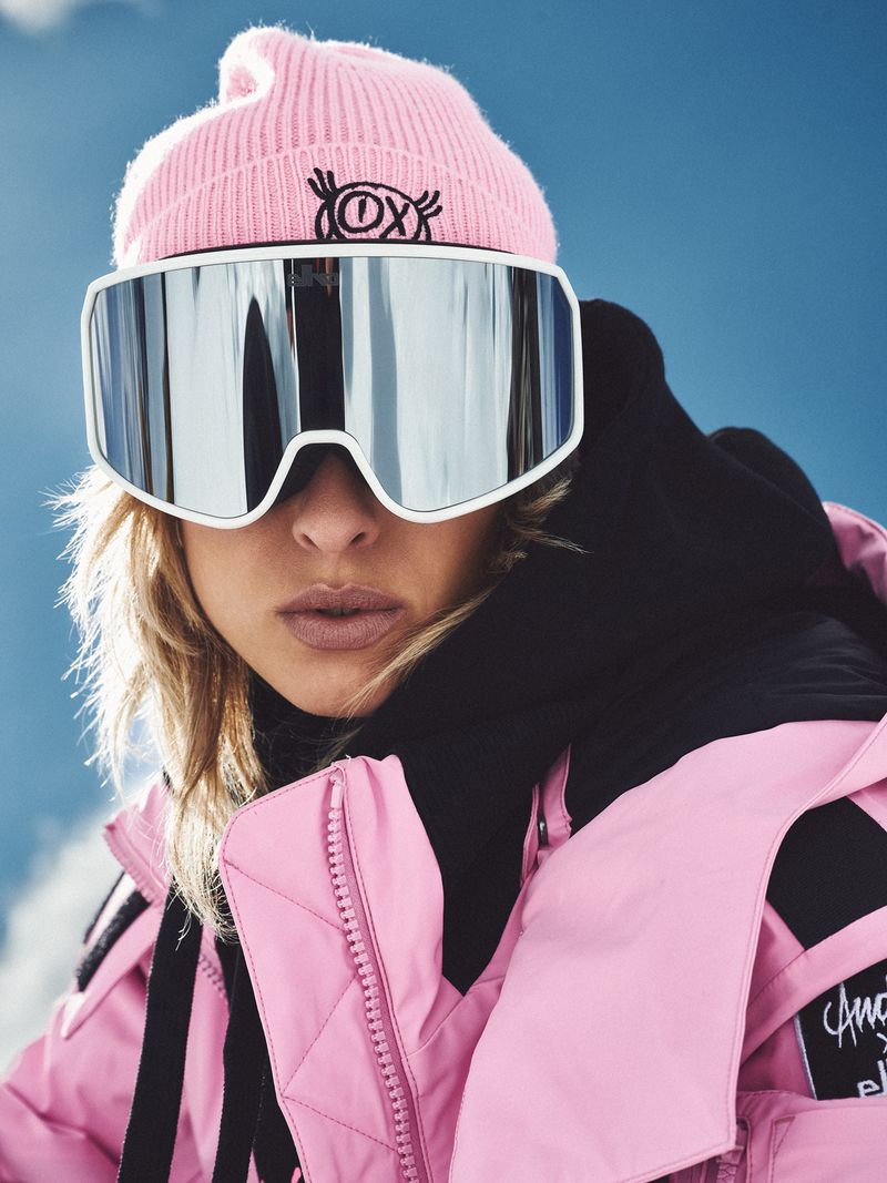 Blond woman in pink beanie and ski jacket wearing oversized ski goggles against a blue sky and snow-covered slope backdrop.