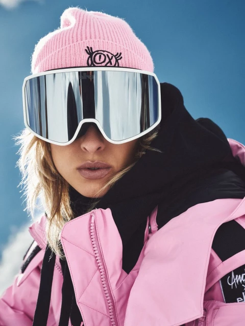 Blond woman in pink beanie and ski jacket wearing oversized ski goggles against a blue sky and snow-covered slope backdrop.