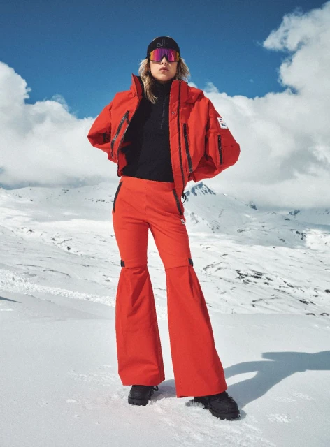 A woman in red ski apparel poses outdoors on a snowy slope against a mountain backdrop under a partly cloudy blue sky.