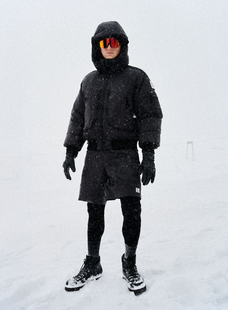 Adult male stands in the snow wearing winter clothing: black puffer coat, black shorts over leggings, gloves, orange glasses and black boots.