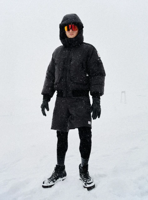 Adult male stands in the snow wearing winter clothing: black puffer coat, black shorts over leggings, gloves, orange glasses and black boots.