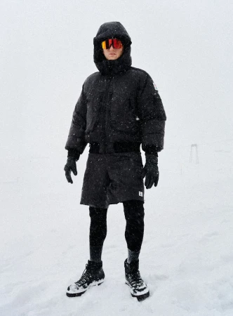 Adult male stands in the snow wearing winter clothing: black puffer coat, black shorts over leggings, gloves, orange glasses and black boots.