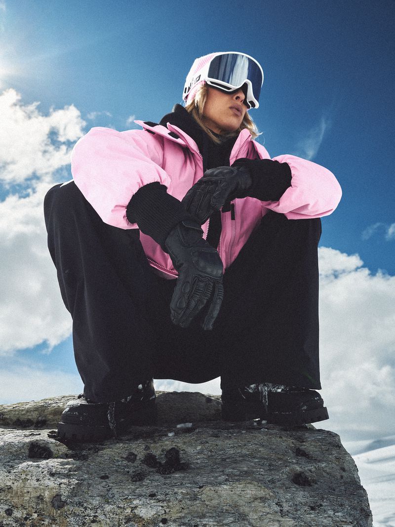 Woman in pink ski jacket, black pants, and goggles sits on a rock in a snowy mountain landscape against a blue sky with clouds.