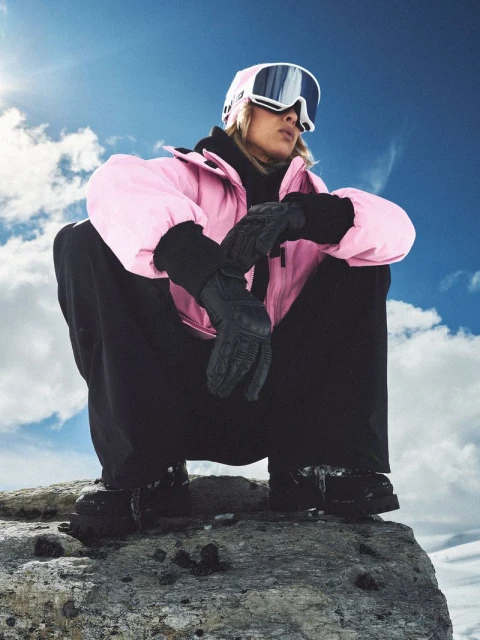 Woman in pink ski jacket, black pants, and goggles sits on a rock in a snowy mountain landscape against a blue sky with clouds.