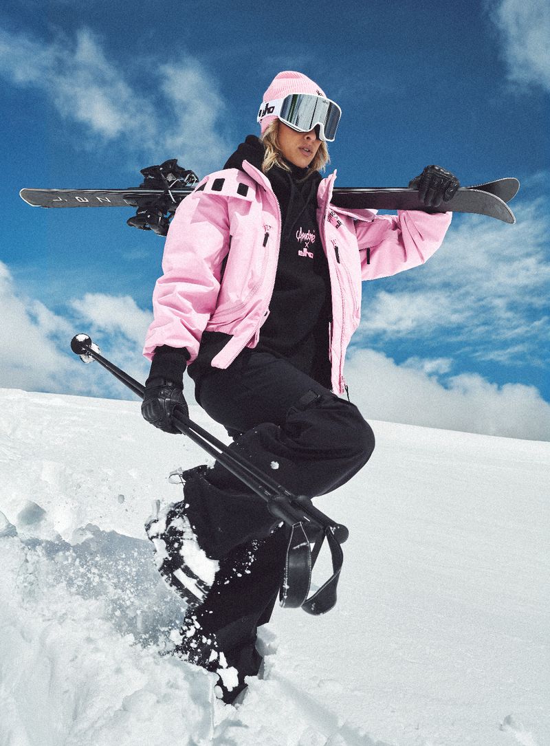 A woman in a pink ski jacket and black snow pants stands on snow with skis and poles, against a snowy hill and a cloudy sky.