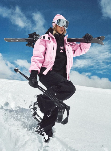 A woman in a pink ski jacket and black snow pants stands on snow with skis and poles, against a snowy hill and a cloudy sky.