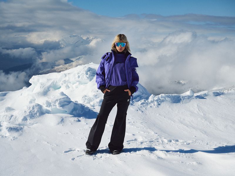 Woman in blue ski jacket and black pants stands on snow covered ground with mountains in background on a sunny day.