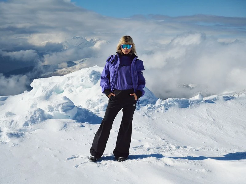 Woman in blue ski jacket and black pants stands on snow covered ground with mountains in background on a sunny day.