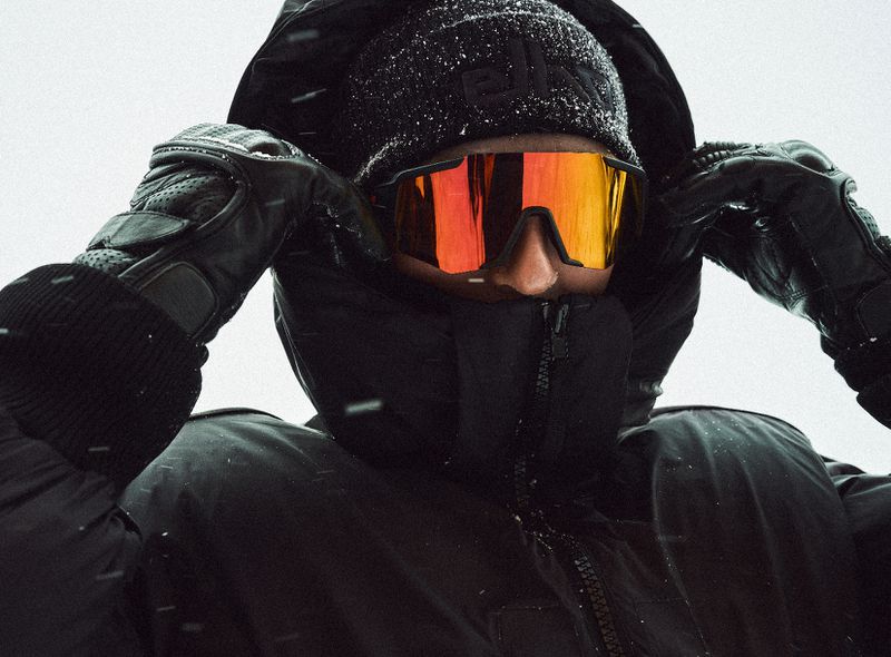 Person in winter clothing, wearing orange-tinted goggles and a black beanie, stands outdoors against a snowy background on a cold day.