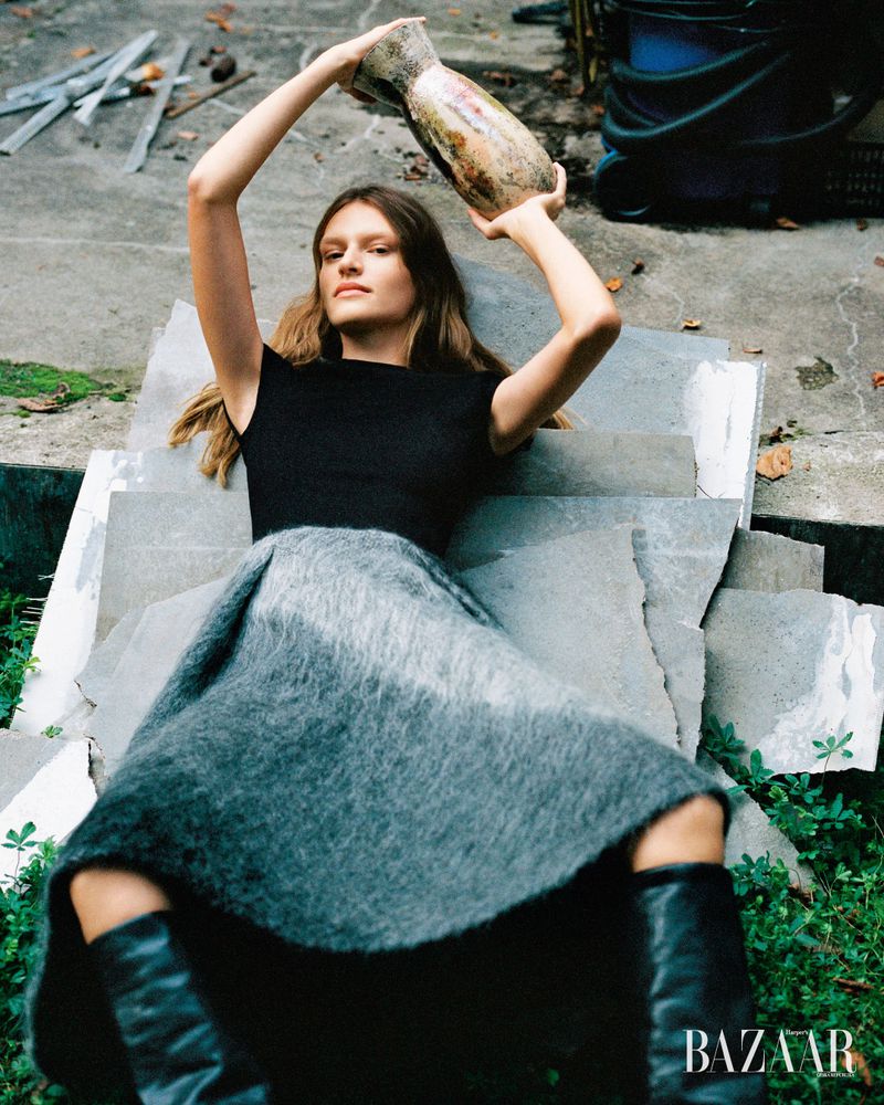 A fashionable woman lies on stone steps holding a vase, wearing a black top, gray skirt, and boots, set against a concrete and plant background.