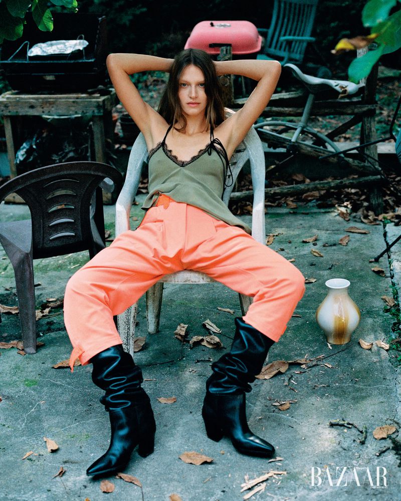 Fashion editorial shot outdoors of a young adult woman sitting in a plastic chair wearing orange pants and black boots with a leafy background.