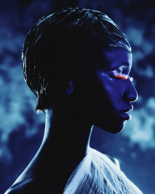 Profile portrait of a woman with wet-look hair and orange makeup on closed eyelids, against a cloudy blue background in studio lighting.
