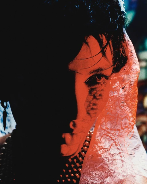 Close-up studio portrait of a woman with fair skin, wearing a pearl necklace and a lace head covering, set against a dark background.