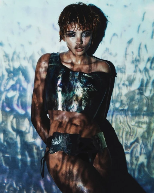 A tanned, muscular adult woman poses indoors wearing an elaborate two-piece metal-like outfit under dynamic blue and white lighting.