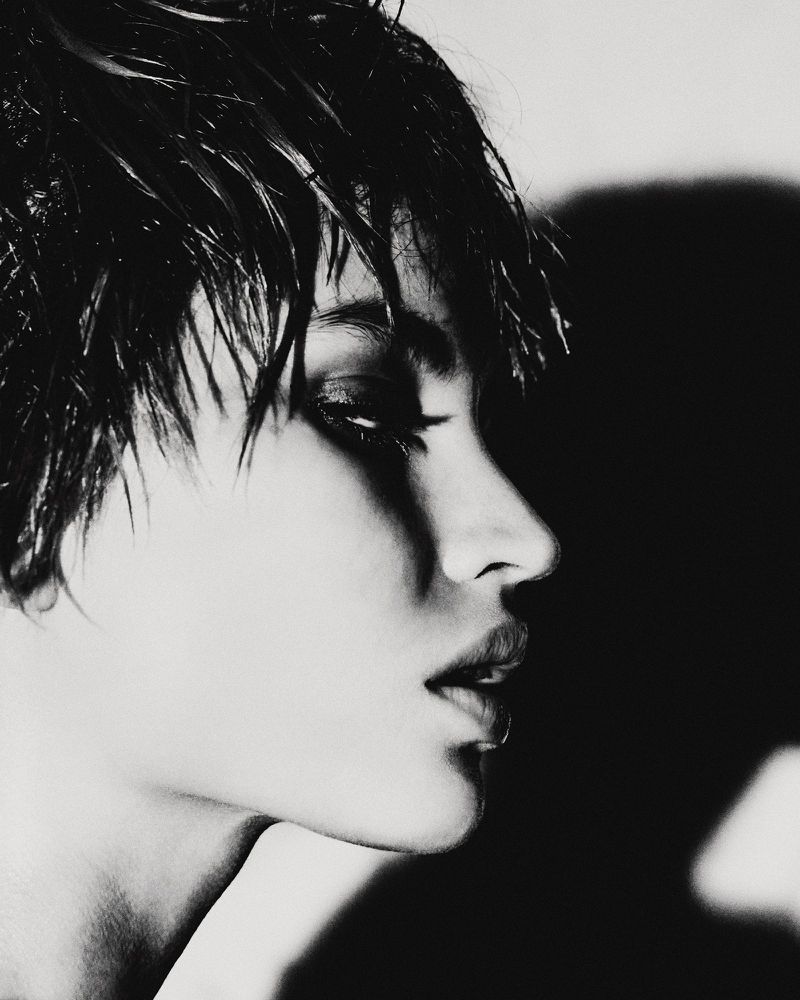 Monochrome portrait of a woman with slicked-back dark hair, side profile view, dramatic lighting, and a neutral expression.