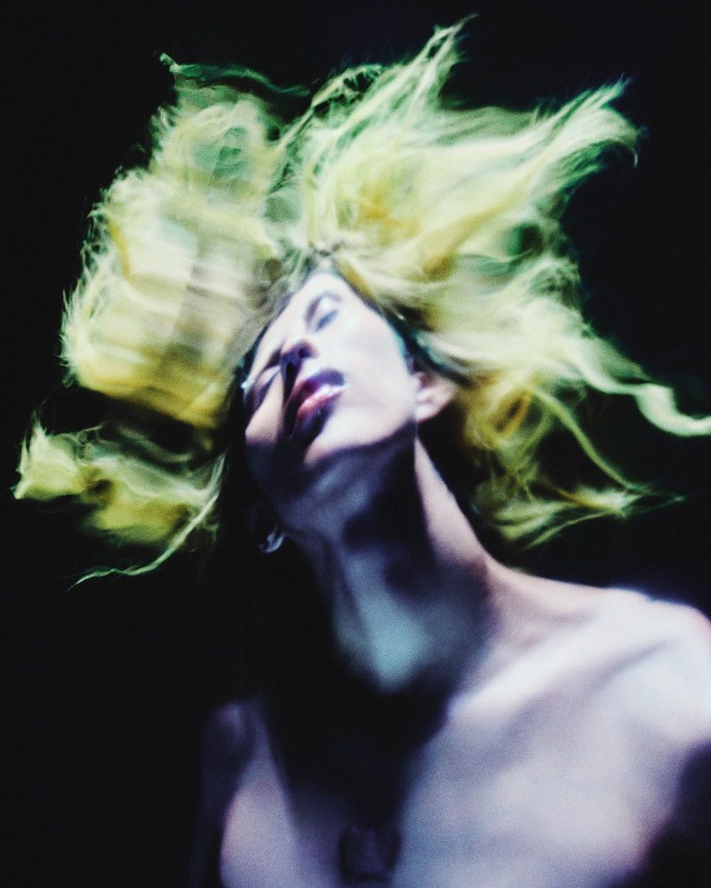 Close-up portrait of a topless person with blonde hair tossed upwards against a dark background, bathed in soft, diffused lighting.
