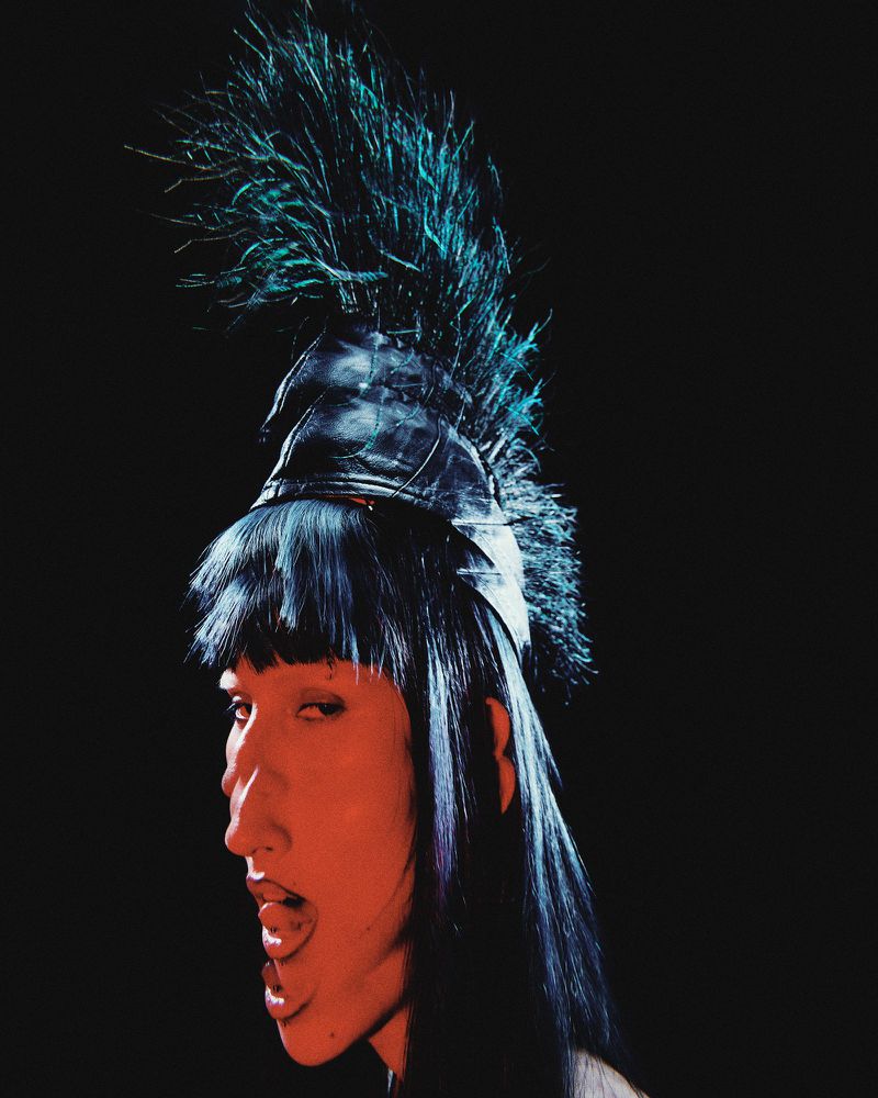Portrait of a person with dark hair and a feather headdress against a black background in a fashion-style image with unique lighting.