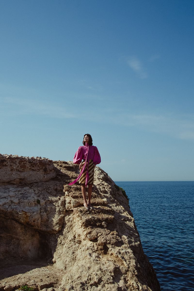 A woman in a pink top and striped skirt stands on a rocky cliff overlooking the ocean under a clear blue sky on a sunny day.