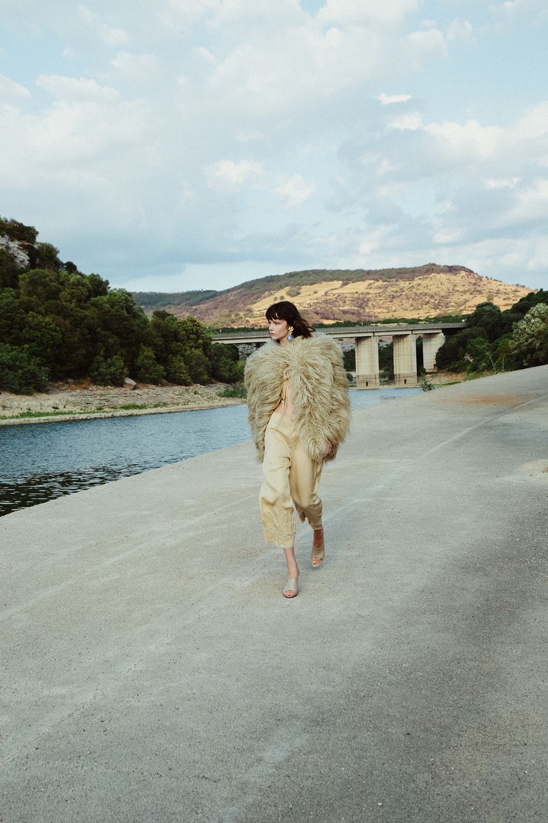 A woman in a furry coat walks along a concrete path by a river under a cloudy sky, with hills and a bridge in the background.